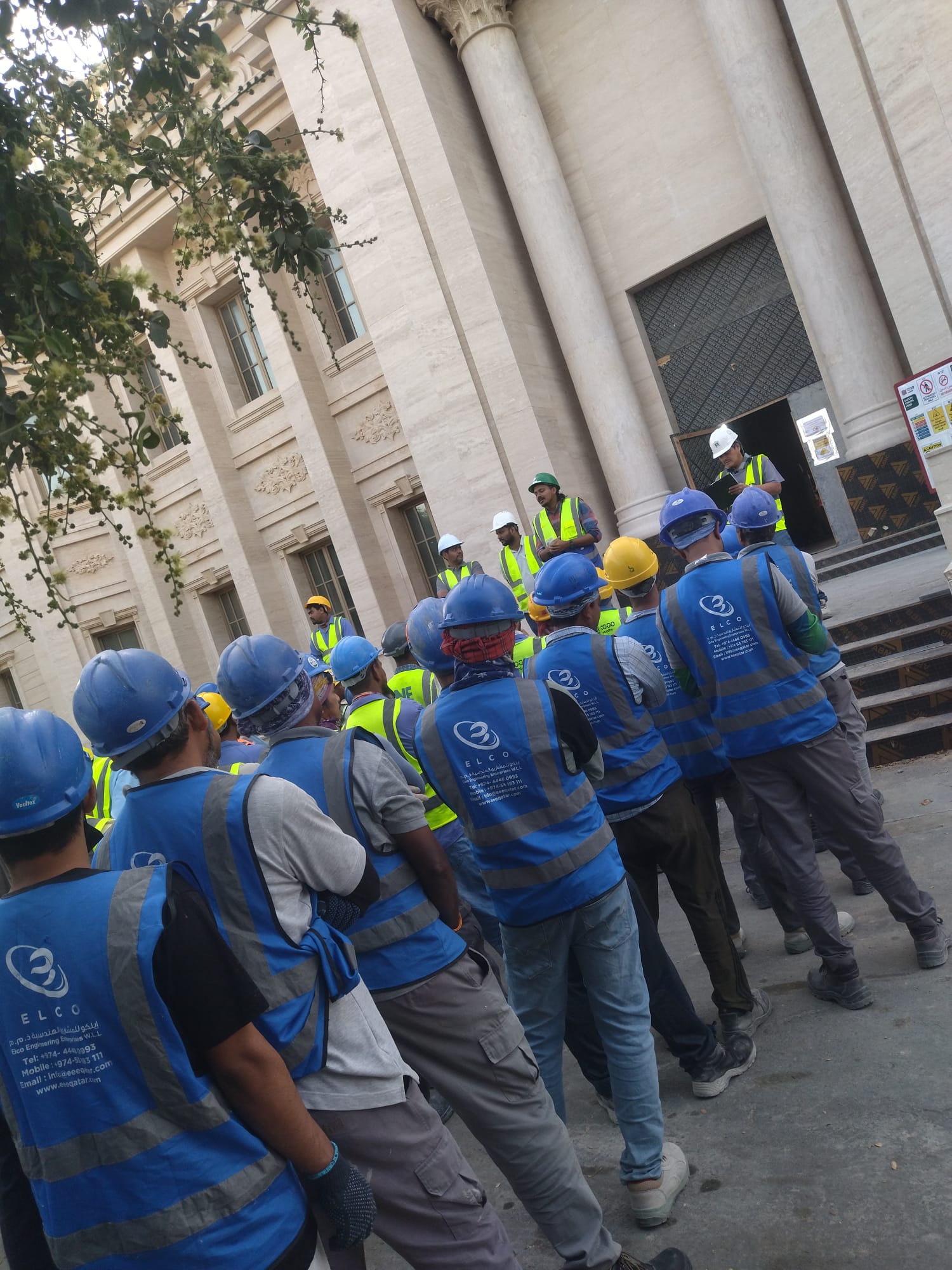 ELCO Engineering team in branded blue vests and helmets gathered at a grand building entrance for site briefing