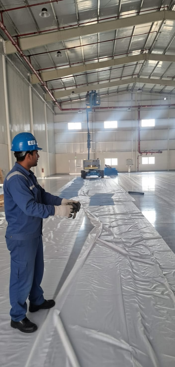 ELCO worker in EEE blue helmet and branded jumpsuit preparing floor protection sheeting in large warehouse with scissor lift in background