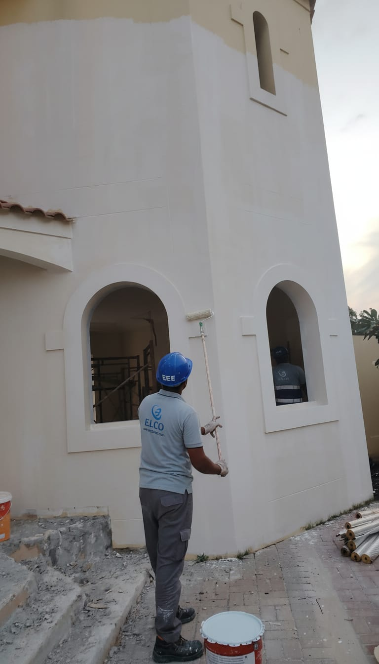 ELCO worker in branded ELCO shirt and EEE blue helmet painting exterior wall of a villa with roller on extension pole