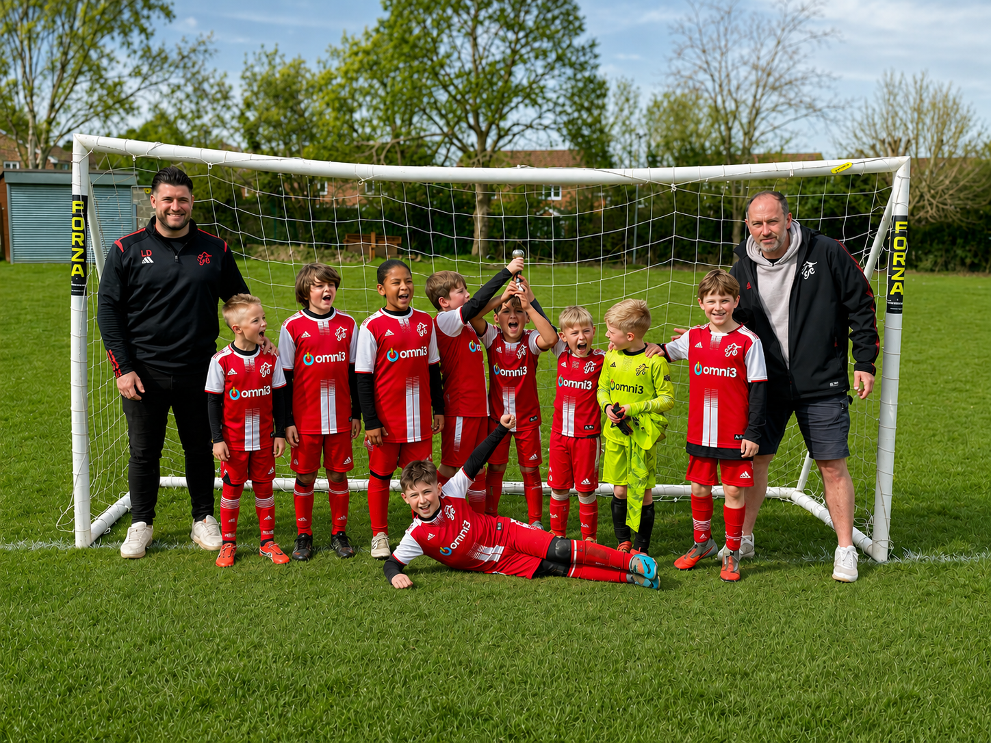 Pulborough Robins Under-9 football squad celebrating in front of a goal in their red and white Adidas kits bearing the Omni3 logo, including a goalkeeper in a yellow kit, with two coaches either side, photographed on a sunny grass pitch in Pulborough, West Sussex.