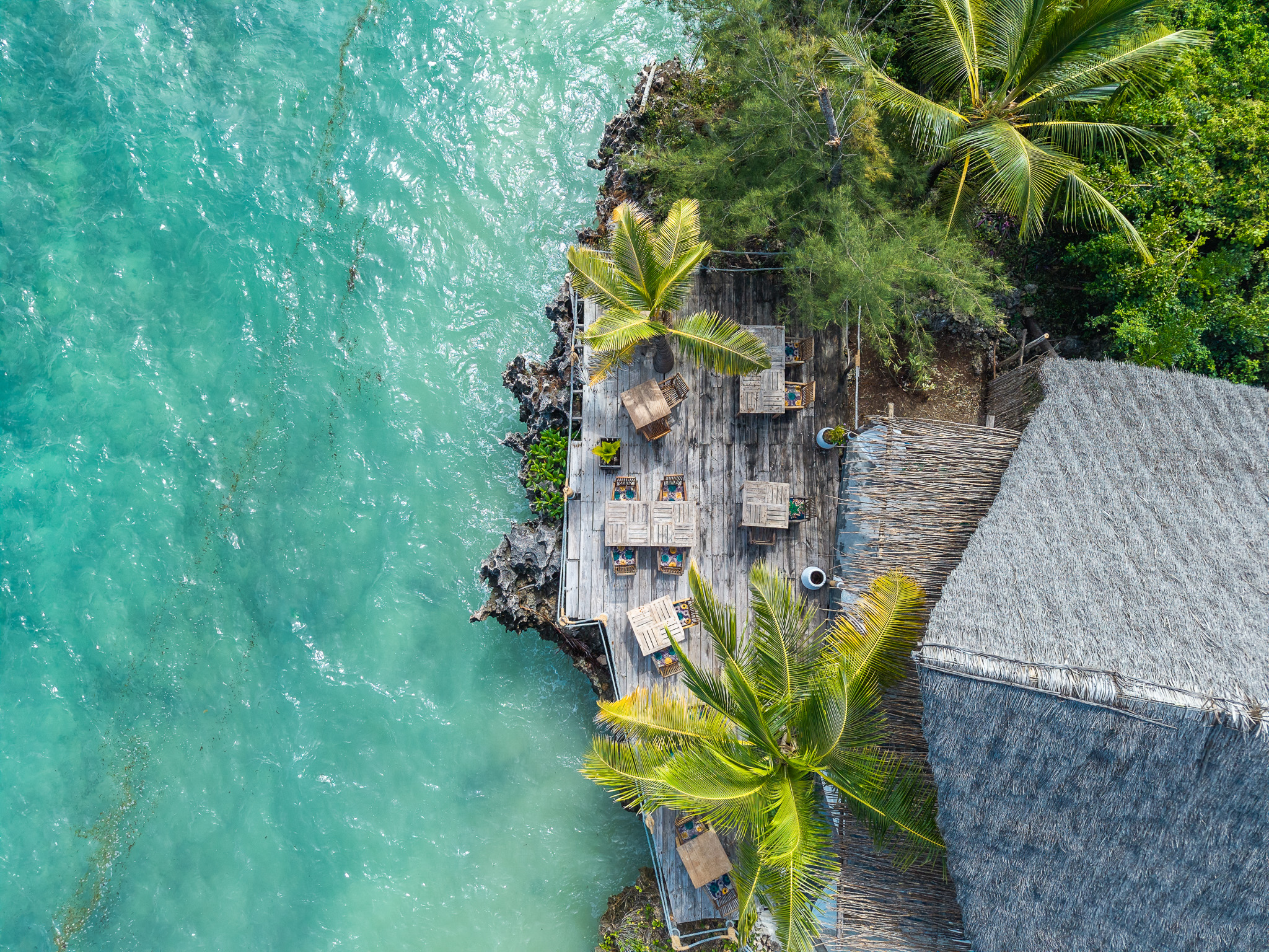Aerial drone view of Tunu Beach restaurant wooden deck over turquoise ocean with palm trees