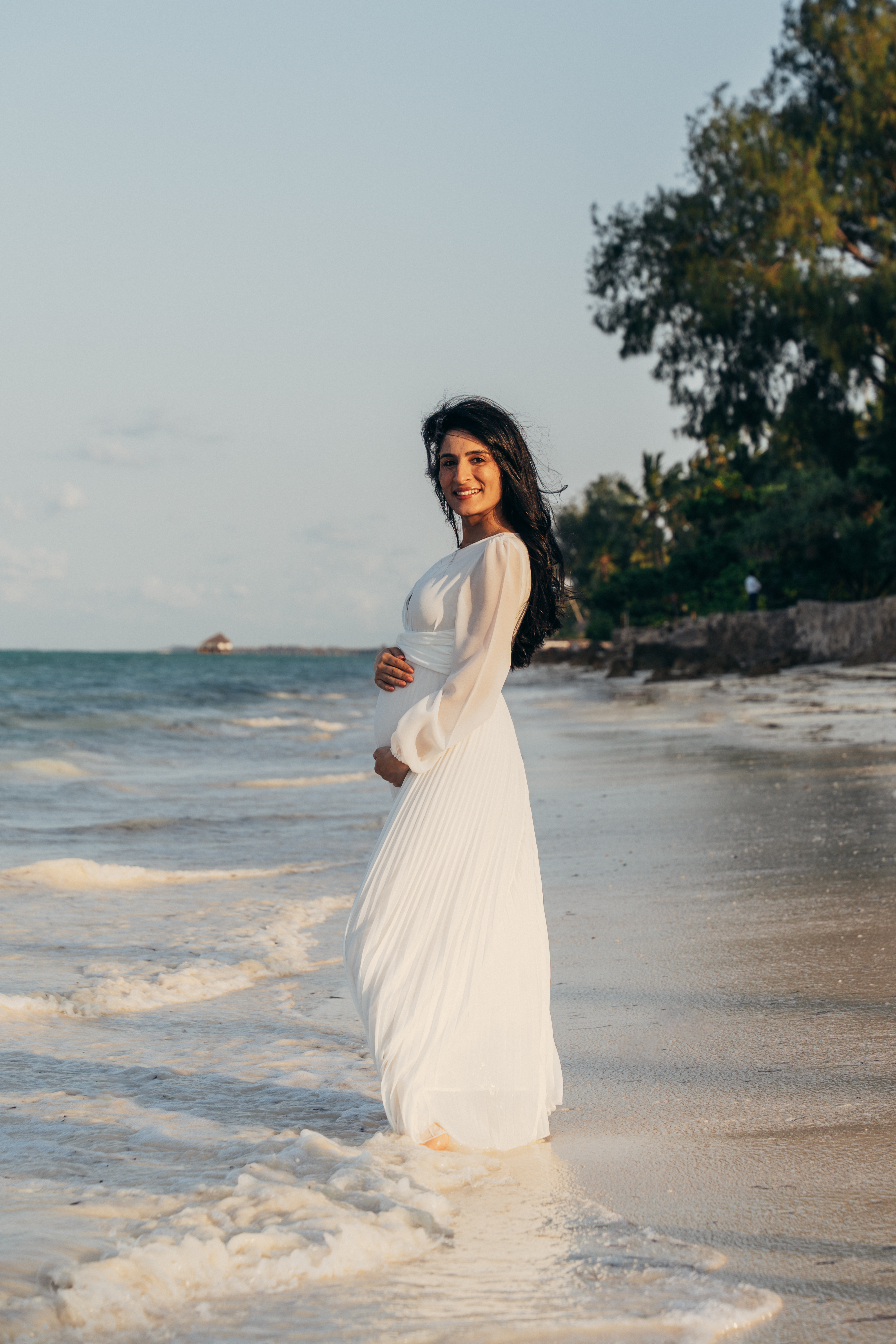 Pregnant woman in white flowing dress standing in ocean waves on Zanzibar beach - maternity portrait