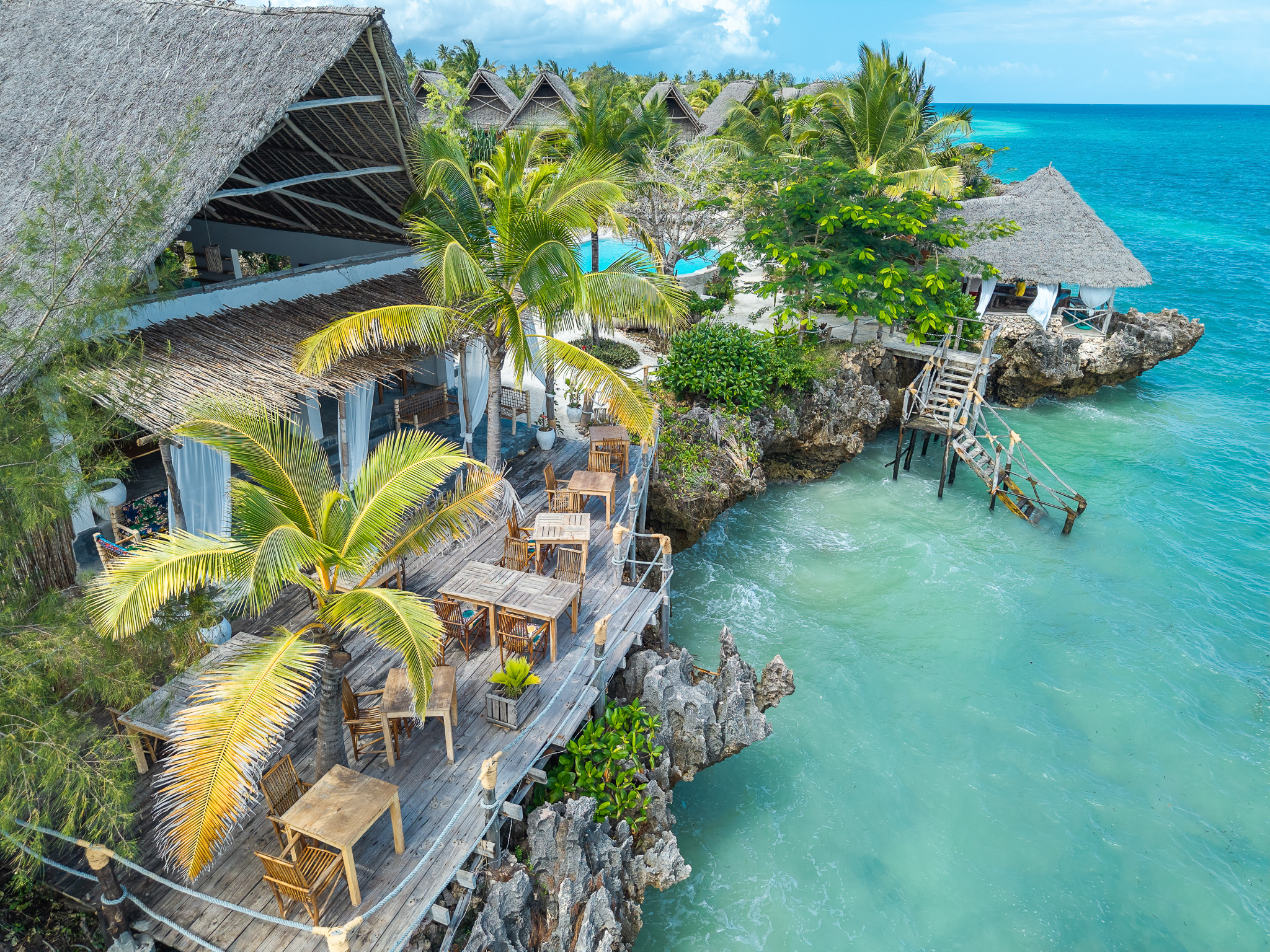 Aerial drone view of Tunu Beach Zanzibar — thatched-roof bungalows, wooden deck restaurant over turquoise Indian Ocean, palm trees and pool