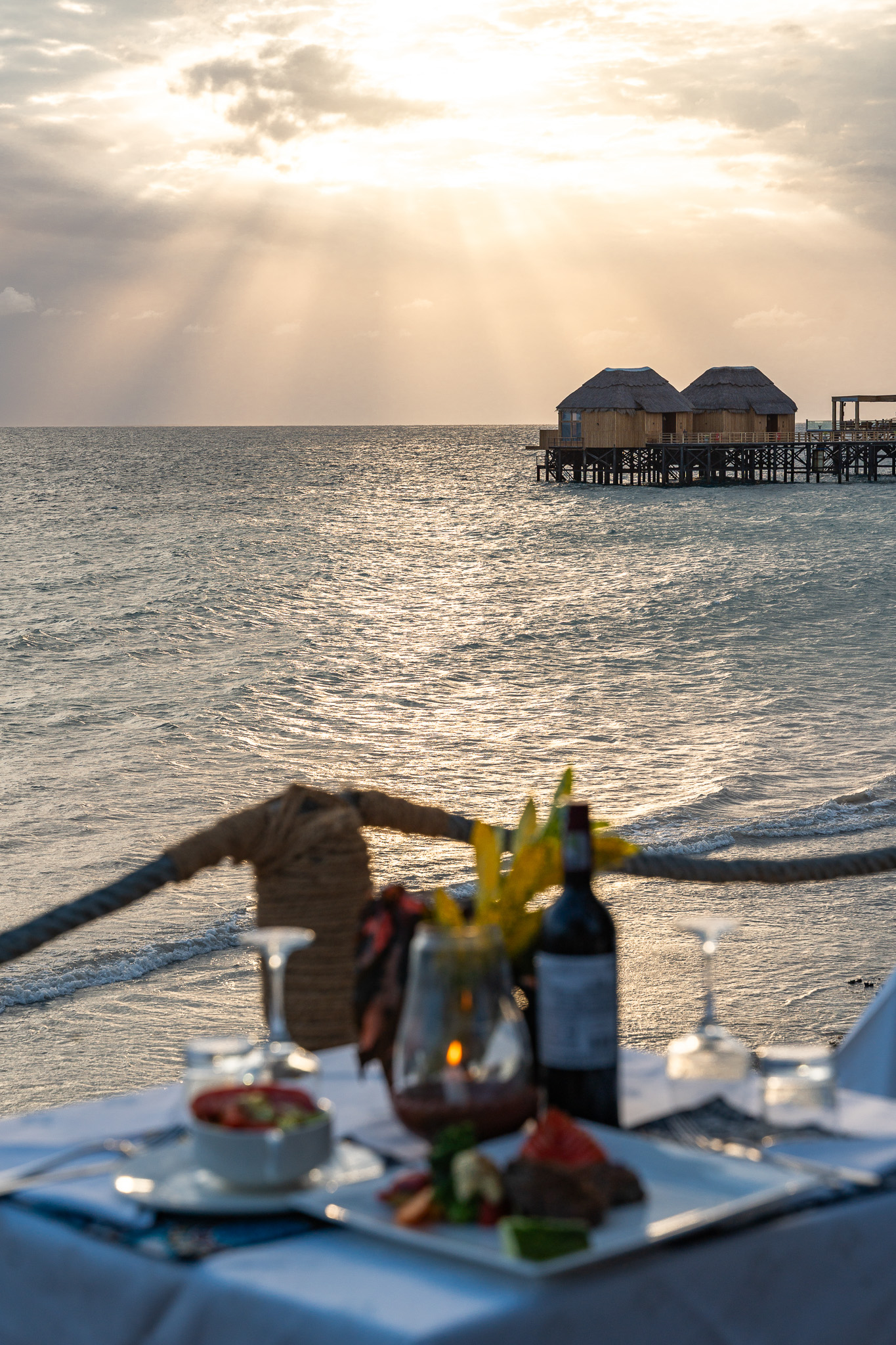 Sunset dinner at Tunu Beach — candlelit table with wine and fresh food, overwater bungalows in golden light